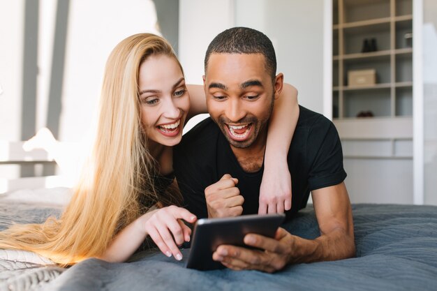 Porträt schöne Momente des glücklichen Paares der niedlichen jungen Frau mit langen blonden Haaren und gutaussehendem Kerl, der im Internet auf Bett in der modernen Wohnung surft. Spaß haben, junge Familie, Liebe