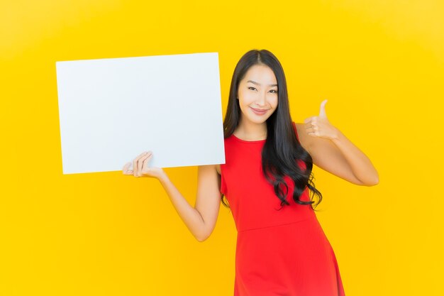 Porträt schönes junges asiatisches Frauenlächeln mit leerer weißer Plakatwand auf gelber Wand