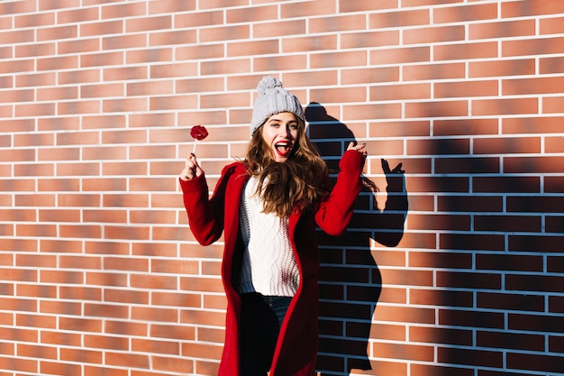 Porträt schönes Mädchen im roten Mantel mit Lutscherlippen an der Wand draußen. Sie trägt eine Strickmütze und sieht zufrieden aus.