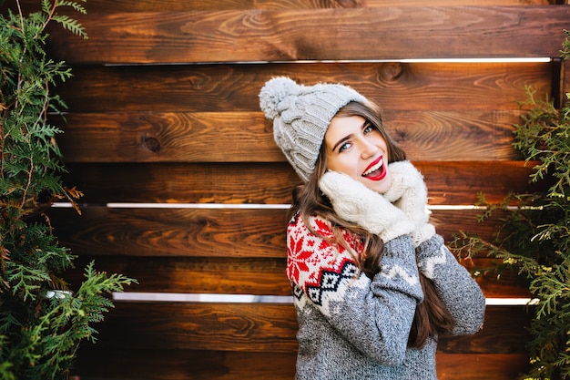 Porträt schönes mädchen mit langen haaren in strickmütze und winterpullover auf holz. sie berührte das gesicht mit händen in handschuhen und lächelte.