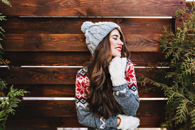 Porträt schönes Mädchen mit langen Haaren und roten Lippen in der warmen Winterkleidung auf Holz. Sie lächelt zur Seite und hält die Augen geschlossen.