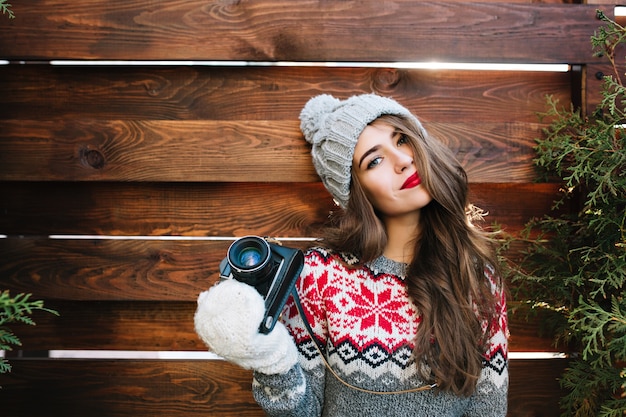 Porträt schönes Mädchen mit roten Lippen in Strickmütze und Handschuhen, die Kamera auf Holz halten.