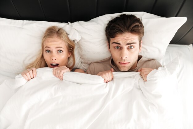 Porträt von den entsetzten Paaren, die im Bett und in der Abdeckung mit Decke liegen