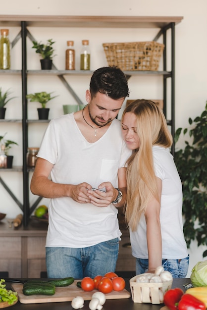 Porträt von den jungen Paaren, die hinter dem Küchentheke betrachtet Mobiltelefon stehen