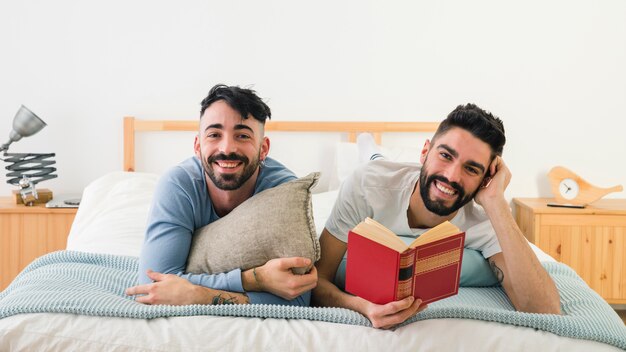 Porträt von den lächelnden jungen homosexuellen Paaren, die auf Front über dem Bett liegen, das Kamera betrachtet