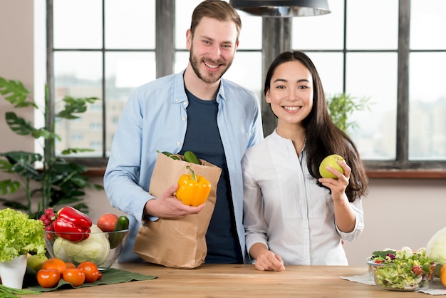 Porträt von den lächelnden jungen Paaren, die hinter der hölzernen Küchenarbeitsplatte hält gelben grünen Pfeffer und grünen Apfel stehen