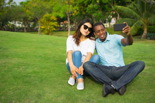 Porträt von den multiethnischen Paaren, die auf Rasen stationieren und selfie mit Smartphone nehmen.