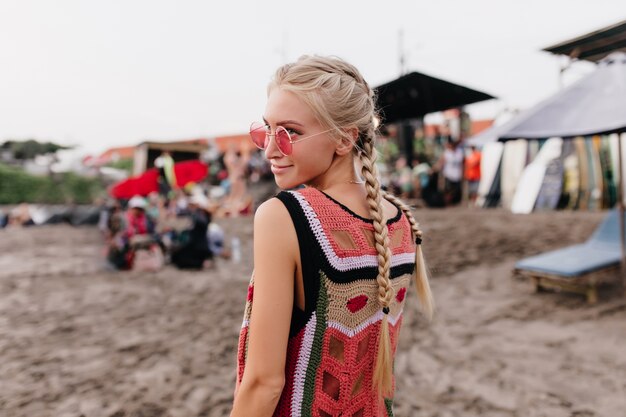 Porträt von der Rückseite der blonden Frau im gestrickten Trägershirt, das mit Lächeln aufwirft. Außenaufnahme der stilvollen Dame mit Zöpfen, die Zeit am Strand verbringen.