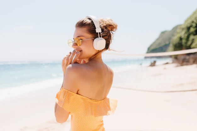 Porträt von der Rückseite der spektakulären Frau, die mit Lächeln an der Ozeanküste aufwirft. Foto im Freien des lachenden wunderbaren Mädchens im orangefarbenen Badeanzug und in den weißen Kopfhörern.