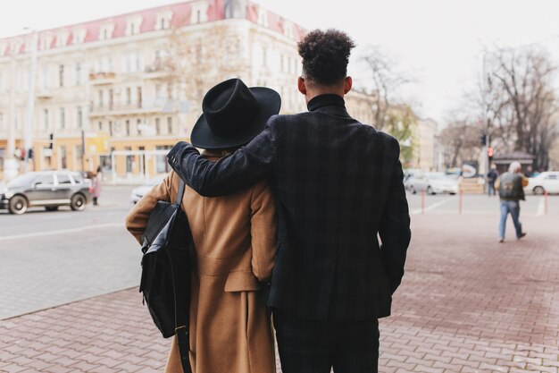 Porträt von der Rückseite des großen Kerls im dunklen Anzug mit dem lockigen Haar, das Timer mit Mädchen im beige Mantel ausgibt. Außenfoto des romantischen Paares, das Stadtansichten genießt.