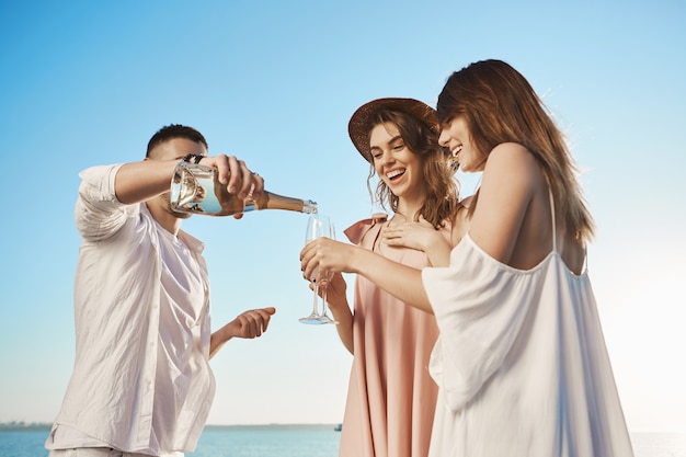 Porträt von drei jungen attraktiven Menschen, die im Urlaub mit der Yacht reisen und Champagner trinken und frische Seeluft genießen. Ein Freund lud zwei Damen zu seinem Boot ein und feierte den Beginn des Sommers.