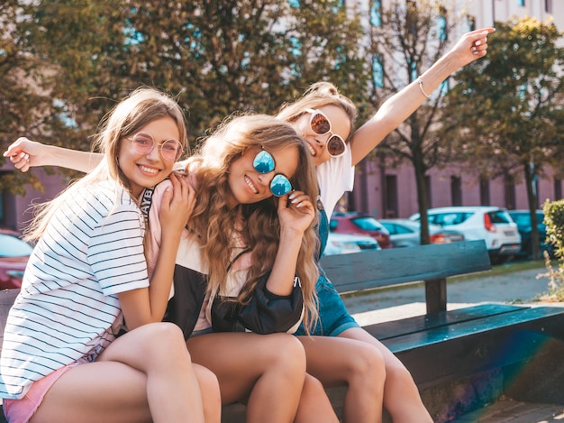 Porträt von drei jungen schönen lächelnden Hippie-Mädchen in der modischen Sommerkleidung Sexy sorglose Frauen, die auf der Bank in der Straße sitzen Positive Modelle, die Spaß in der Sonnenbrille haben Hände anheben