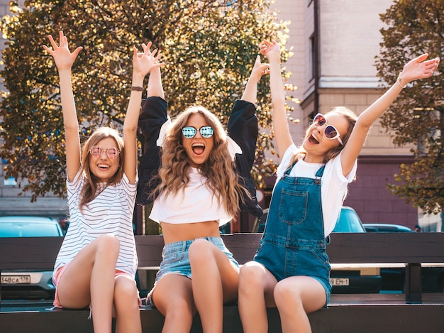 Porträt von drei jungen schönen lächelnden Hippie-Mädchen in der modischen Sommerkleidung Sexy sorglose Frauen, die auf der Bank in der Straße sitzen Positive Modelle, die Spaß in der Sonnenbrille haben Hände anheben