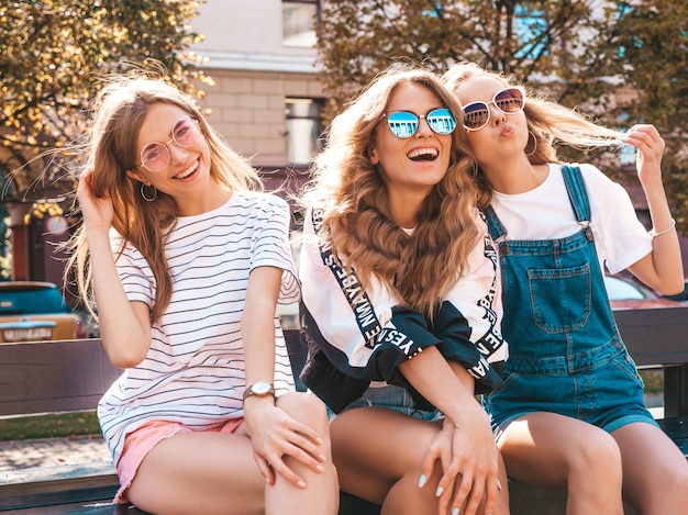 Porträt von drei jungen schönen lächelnden Hippie-Mädchen in der modischen Sommerkleidung Sexy sorglose Frauen, die auf der Bank in der Straße sitzen Positive Modelle, die Spaß in der Sonnenbrille haben