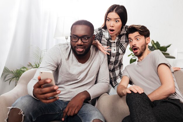 Porträt von drei schockierten Freunden, die zu Hause auf Stühlen sitzen und erstaunt ins Handy schauen, während sie isoliert Zeit miteinander verbringen