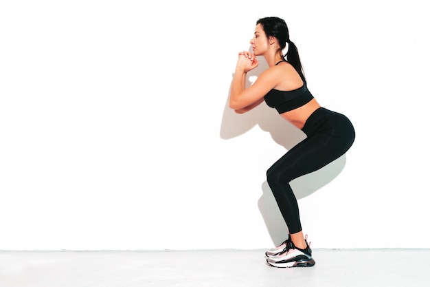 Porträt von Fitness selbstbewusste Frau in schwarzer Sportkleidung Sexy junges schönes Modell mit perfektem Körper Frau isoliert auf weißer Wand im Studio Stretching vor dem Training Kniebeugen machen