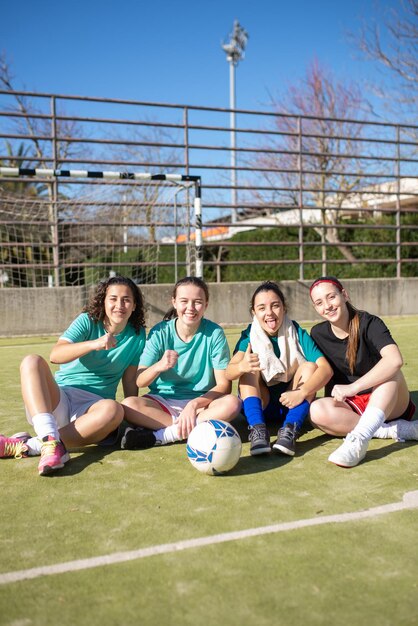 Porträt von Fußballmädchen, die sich während der Erholung amüsieren. Ein Team von vier glücklichen Mädchen sitzt entspannt auf dem Boden und ruht sich nach dem Spiel aus und demonstriert ihre Stärke für die Kamera. Mannschaftssport und aktives Erholungskonzept