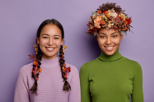 Porträt von glücklichen Mischlingsfrauen in warmen Kleidern, mit Herbstlaub verziertem Haar, drücken gute Gefühle aus, stehen nebeneinander.