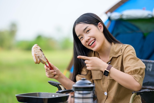 Porträt von Porträt einer glücklichen jungen asiatischen Frau, die allein gegrilltes Schweinefleisch in der Picknickpfanne campt und Essen kocht, während sie auf einem Stuhl auf dem Campingplatz sitzt