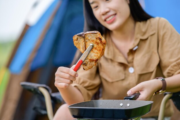 Porträt von Porträt einer glücklichen jungen asiatischen Frau, die allein gegrilltes Schweinefleisch in der Picknickpfanne campt und Essen kocht, während sie auf einem Stuhl auf dem Campingplatz sitzt