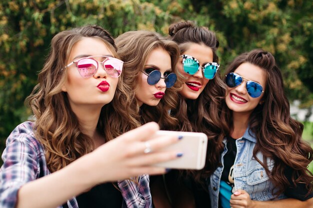 Porträt von vier Freundinnen, die lässige, trendige Kleidung, Frisuren und Make-up tragen, posieren, während sie am Tag draußen im Park ein Selbstporträt per Handy machen.