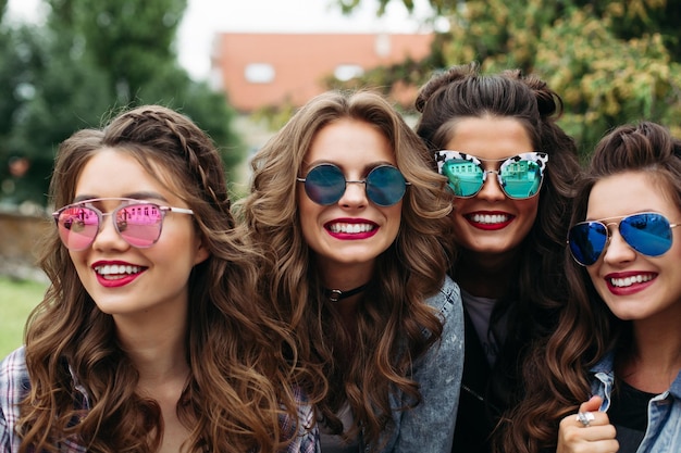 Porträt von vier Freundinnen, die lässige, trendige Kleidung, Frisuren und Make-up tragen, posieren, während sie am Tag draußen im Park ein Selbstporträt per Handy machen.