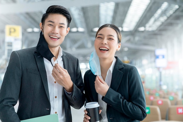 Porträt von zwei asiatischen Geschäftsleuten tragen ein virenschützendes Lächeln mit Gesichtsmasken und einem willkommenen und selbstbewussten Blick in die Kamera mit unscharfem Hintergrund des Flughafenterminals, der den neuen normalen Lebensstil sozial distanziert