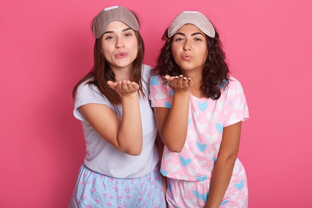 Porträt von zwei Frauen, die Hände heben, Schlagkuss senden, ins Bett gehen, Pyjama und Schlafmasken tragen