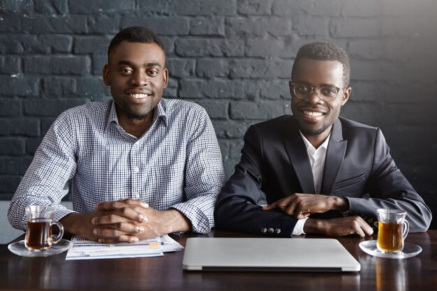 Porträt von zwei gutaussehenden selbstbewussten afroamerikanischen Personalvermittlern, die am Schreibtisch dicht beieinander sitzen