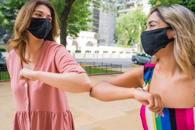 Porträt von zwei jungen Freunden, die Gesichtsmaske tragen und Ellbogen im Freien stoßen.