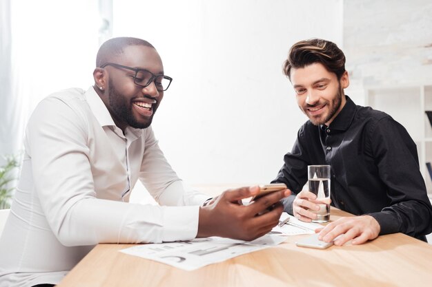 Porträt von zwei jungen fröhlichen multinationalen Geschäftsleuten, die am Tisch sitzen und sich glücklich unterhalten, während sie im isolierten Büro zusammenarbeiten