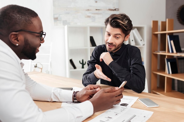 Porträt von zwei jungen lächelnden multinationalen Geschäftsleuten, die am Tisch sitzen und glücklich über neue Projekte diskutieren, während sie isoliert im Büro zusammenarbeiten