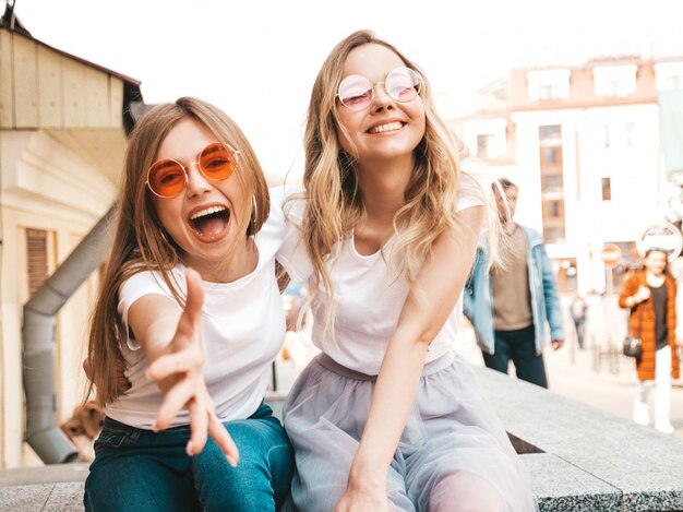 Porträt von zwei jungen schönen blonden lächelnden Hippie-Mädchen im weißen T-Shirt des modischen Sommers kleidet. Sexy sorglose Frauen, die auf Straße sitzen. Positive Modelle, die Spaß in der Sonnenbrille haben