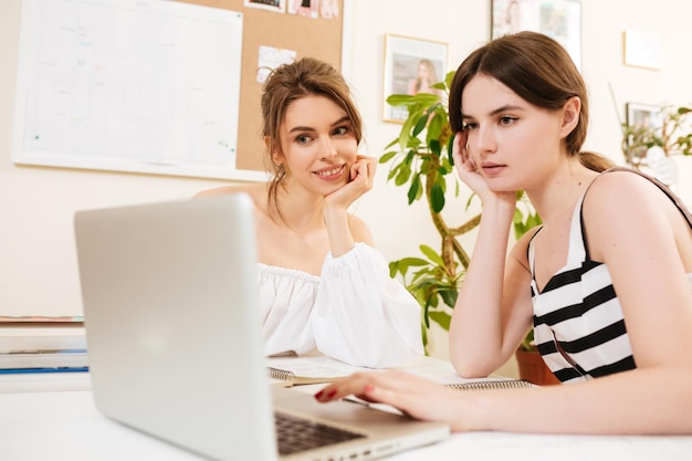 Porträt von zwei jungen schönen Damen, die am Schreibtisch sitzen und zusammenarbeiten, während sie nachdenklich im Laptop im Büro suchen