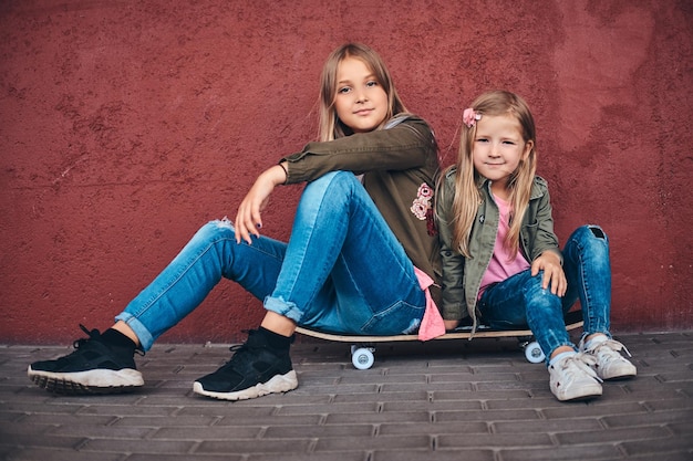 Kostenloses Foto porträt von zwei kleinen schwestern in trendigen kleidern, die sich an eine wand lehnen, während sie auf einem skateboard am brückenfußweg sitzen.
