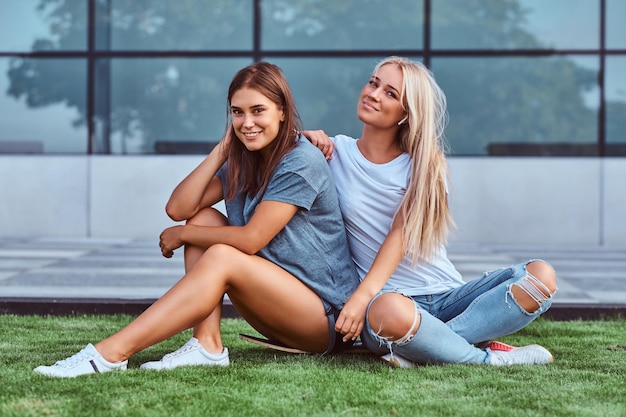 Porträt von zwei lächelnden Mädchen, die auf einem Skateboard auf einem Rasen vor dem Hintergrund des Wolkenkratzers sitzen.