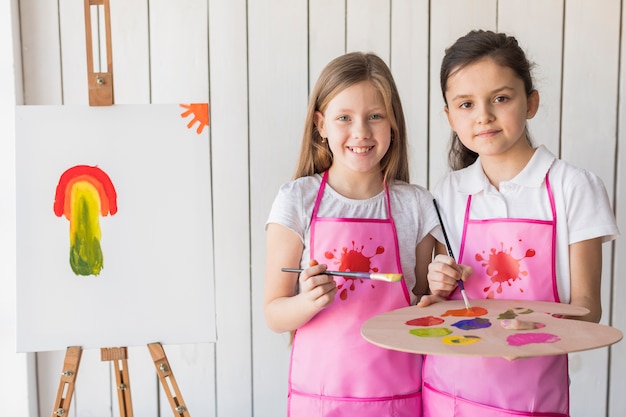 Porträt von zwei lächelnden Mädchen im rosa Schutzblech, das Kamera beim Malen auf dem Gestell betrachtet