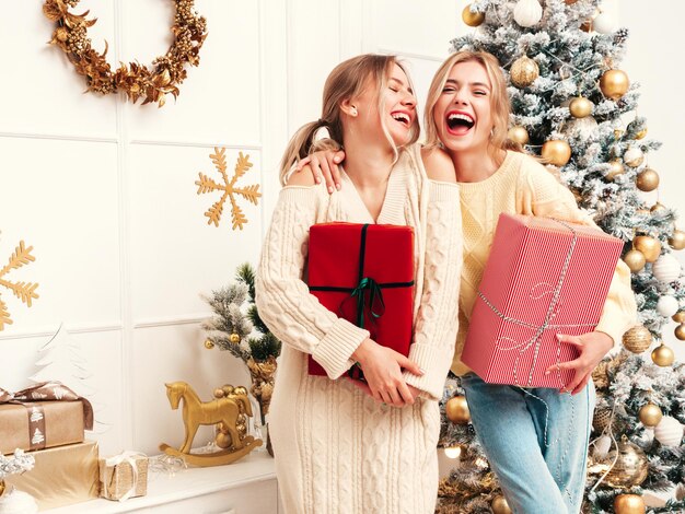 Porträt von zwei schönen blonden Frauen Models posieren in der Nähe von geschmücktem Weihnachtsbaum zu Silvester Frauen, die Spaß haben, bereit zum Feiern Freunde, die in warmen Winterpullovern gekleidet sind