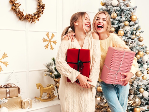 Porträt von zwei schönen blonden Frauen Models posieren in der Nähe von geschmücktem Weihnachtsbaum zu Silvester Frauen, die Spaß haben, bereit zum Feiern Freunde, die in warmen Winterpullovern gekleidet sind