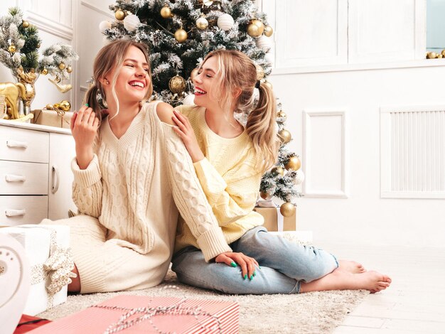 Porträt von zwei schönen blonden Frauen Models posieren in der Nähe von geschmücktem Weihnachtsbaum zu Silvester Frauen, die Spaß haben, bereit zum Feiern Freunde, die in warmen Winterpullovern gekleidet sind