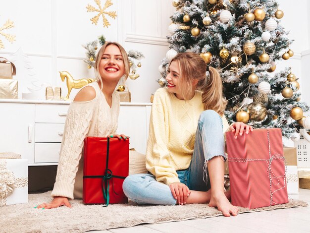 Porträt von zwei schönen blonden Frauen Models posieren in der Nähe von geschmücktem Weihnachtsbaum zu Silvester Frauen, die Spaß haben, bereit zum Feiern Freunde, die in warmen Winterpullovern gekleidet sind