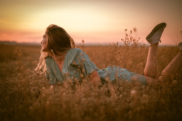 Porträtaufnahme einer glücklichen kaukasischen blonden Frau in einem Sommerkleid, die während des Sonnenuntergangs auf einem Feld liegt