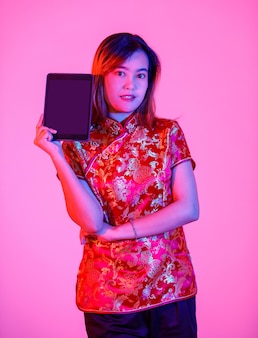 Porträtstudioaufnahme des asiatischen jungen jugendlich weiblichen modells im schönen roten chinesischen traditionellen cheongsam-qipao-hemd, das mit leerer monitor-touchscreen-tablette auf rosa hellem hintergrund steht.