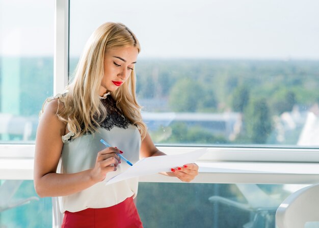 Portrait der blonden jungen Geschäftsfrau, die Dokument nahe dem Fenster überprüft