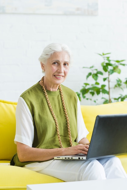 Portrait der lächelnden jungen Frau, die auf gelbem Sofa mit Laptop sitzt