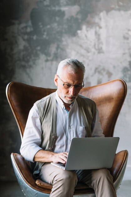 Portrait des älteren Mannes sitzend auf Stuhl unter Verwendung des Laptops