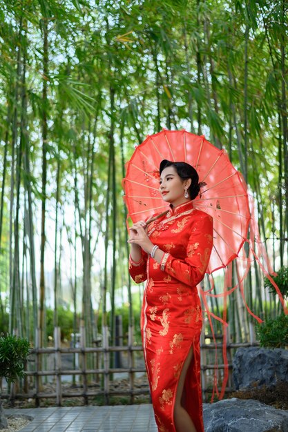 Portrait hübsche asiatische Frau in einem chinesischen Cheongsam posiert mit schönem rotem Papierschirm auf Bambuswald, Kopierraum