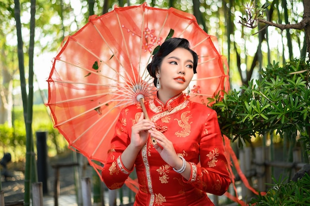 Portrait hübsche asiatische Frau in einem chinesischen Cheongsam posiert mit schönem rotem Papierschirm auf Bambuswald, Kopierraum