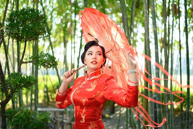 Kostenloses Foto portrait hübsche asiatische frau in einem chinesischen cheongsam posiert mit schönem rotem papierschirm auf bambuswald