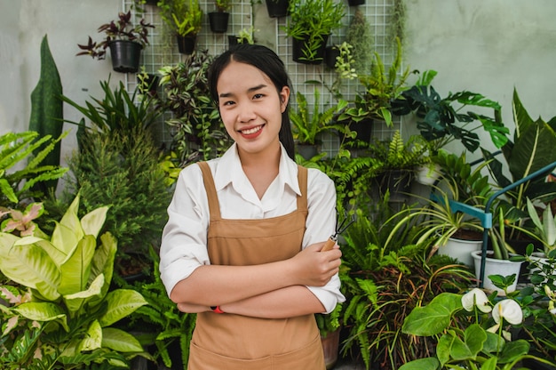 Portrait junge asiatische Gärtnerin mit Schürzenständer und Kreuzarm, sie lächelt und schaut zur Kamera